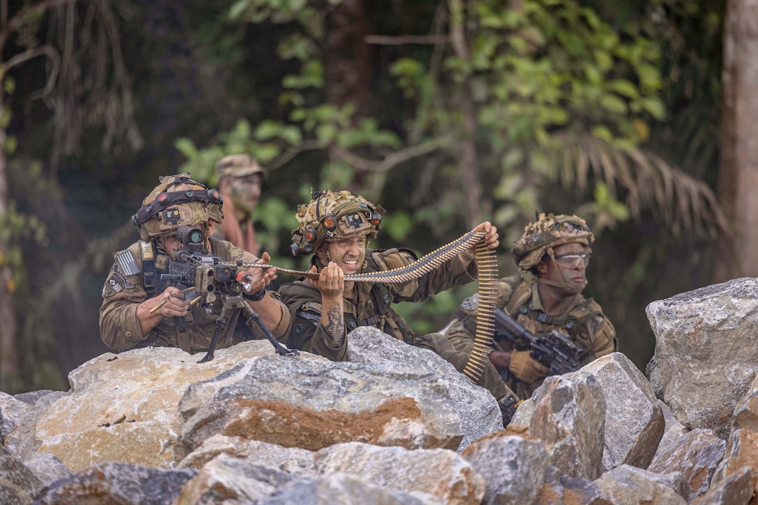 Three soldiers kneel behind rocks as one fires a weapon while another holds ammo and another looks in the distance with greenery in the background.