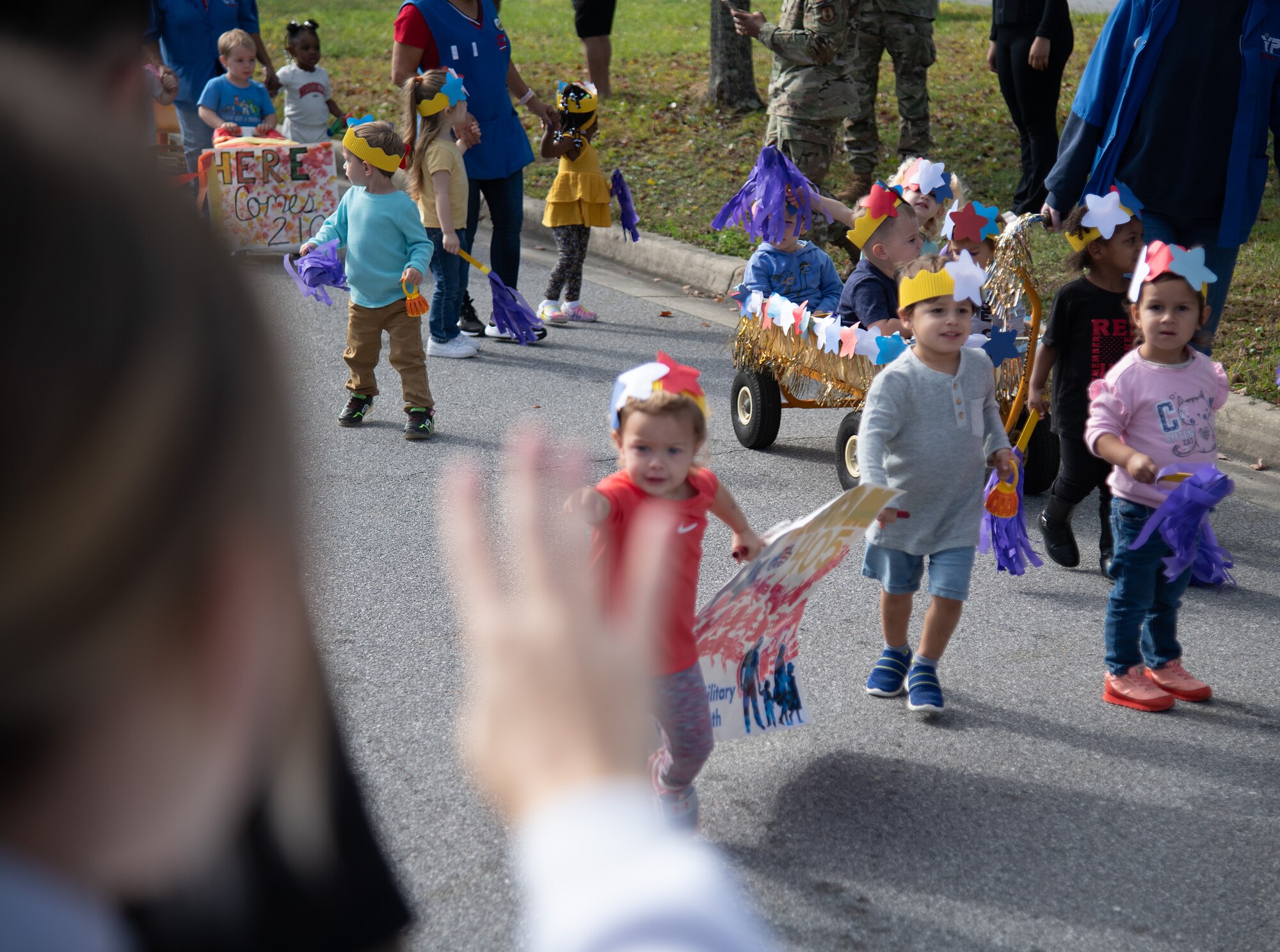 A person waves in the foreground at a group of toddlers and floats of babies at a parade.
