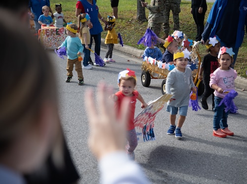 A person waves in the foreground at a group of toddlers and floats of babies at a parade.