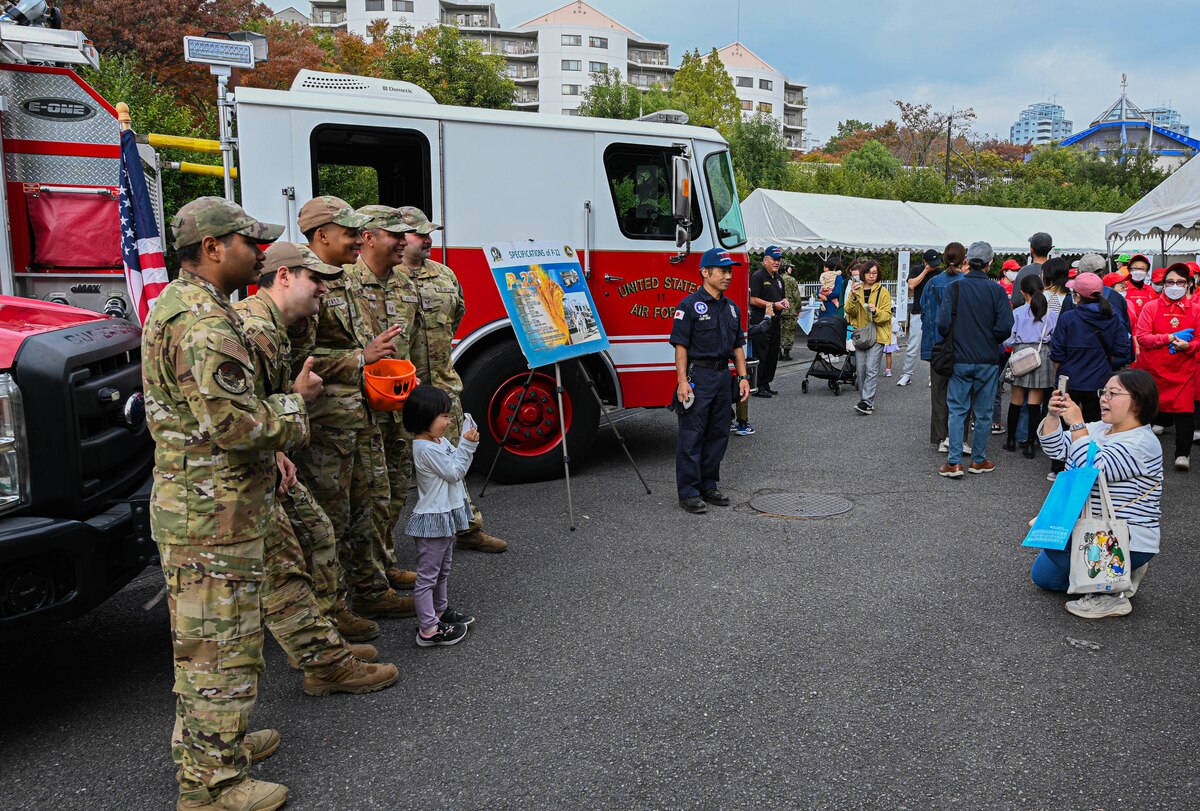5 men in uniform pose for photo with fire trucks parked behind them.