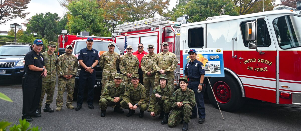 14 people in uniforms pose for a photo Infront of 3 fire trucks.