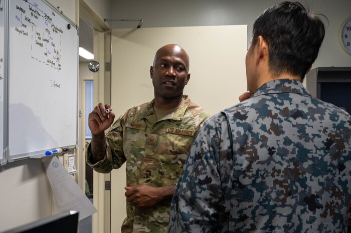 Two people in uniform are in the photo. They are talking to each other while one is standing by a white board with writings on it.,