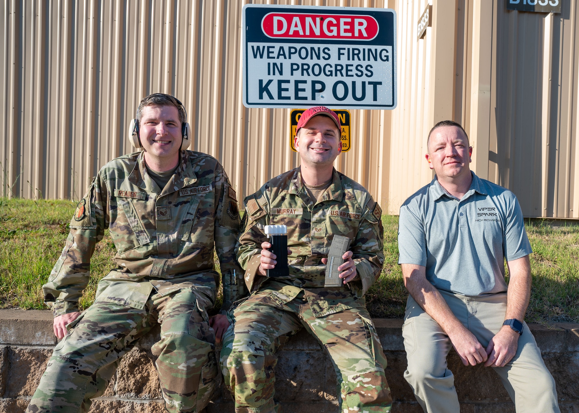 Three men sit on a ledge, the man in the center is holding a 3-D printed magazine loader and magazine for the M-4 Carbine.