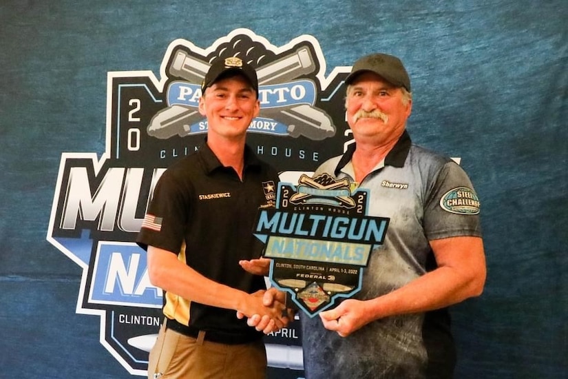 Two men standing together while shaking hands. One man is holding a plaque for winning a shooting competition.