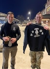 Two men pose together on a sidewalk, one man is holding an Army Airborne sweatshirt and the other man is holding a water bottle and a hat.