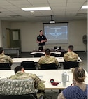 Man in Army polo stands at the front of a classroom giving a presentation to people in Army uniforms.