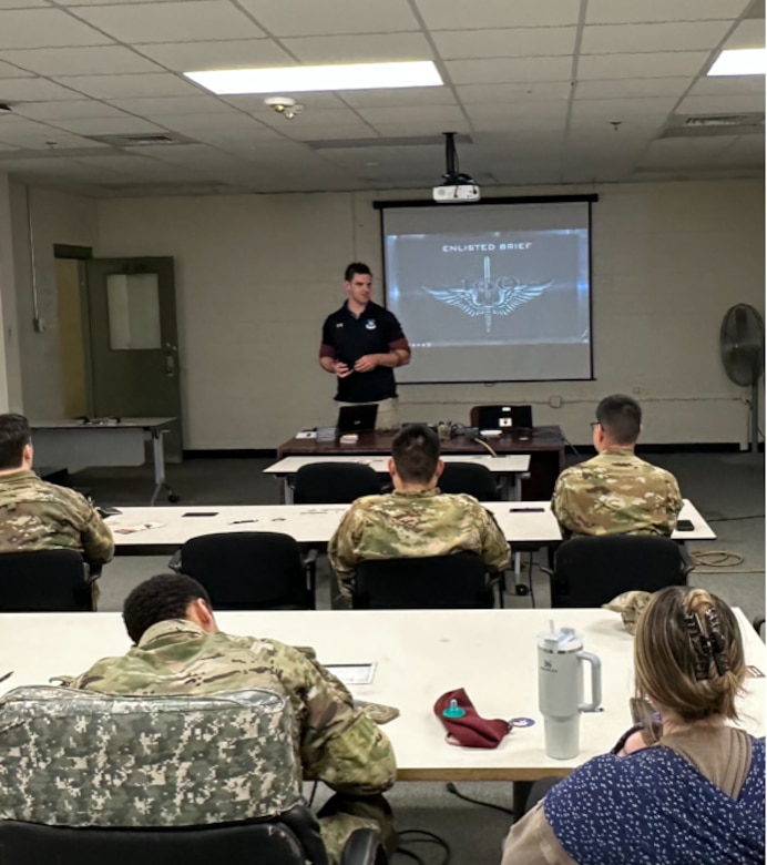 Man in Army polo stands at the front of a classroom giving a presentation to people in Army uniforms.