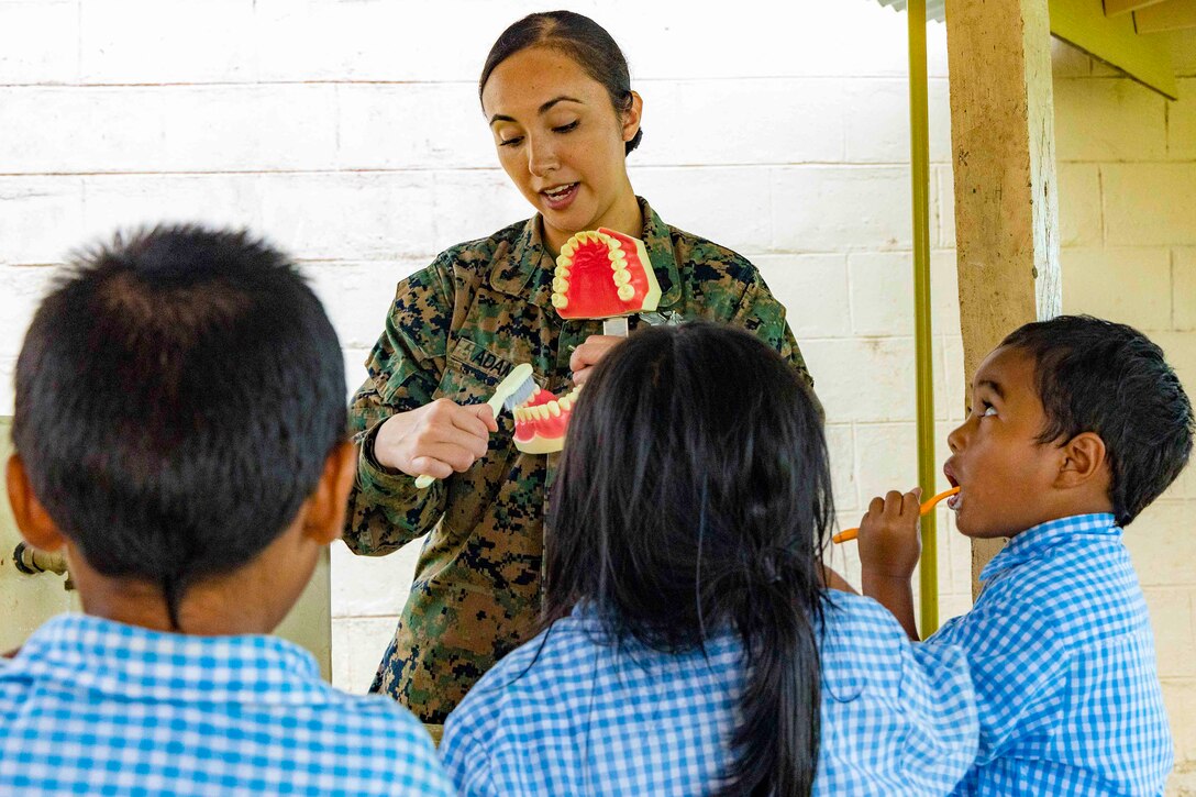 A sailor brushes the teeth on a plastic model of a set of teeth in front of three children, one of whom brushes their own teeth.
