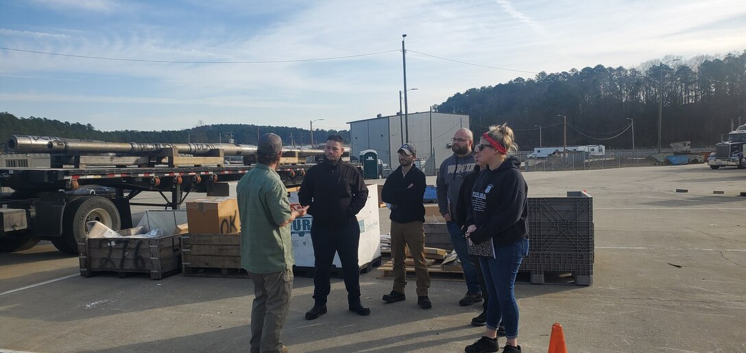Group of people standing in a concrete yard with boxes and a flatbed truck
