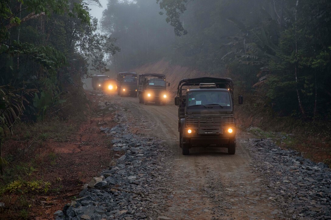 Vehicle headlights illuminate a trail as they travel a dirt road in a forest under a foggy sky.