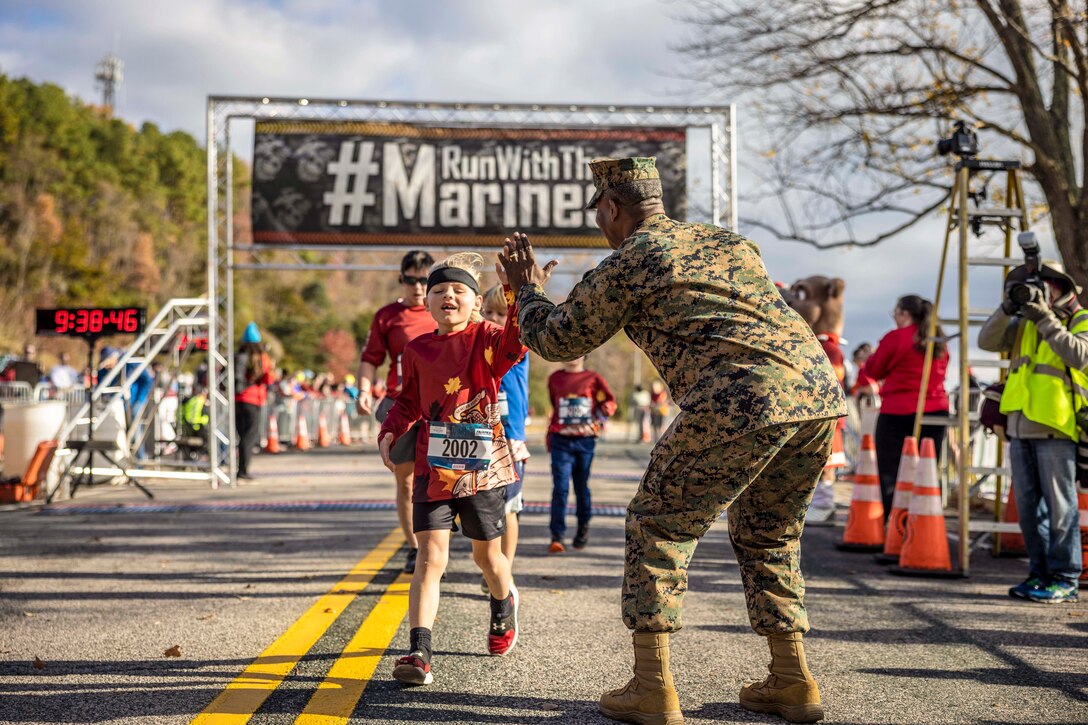 A Marine high-fives a military child after a run as a photographer takes their picture while fellow participants run across the finish line in the background.
