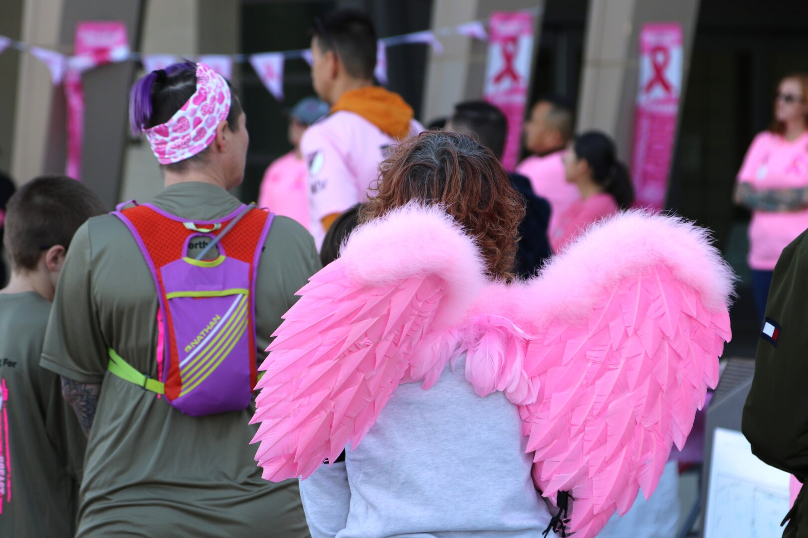 Back view of woman wearing pink angel wings standing next to another woman with headband.