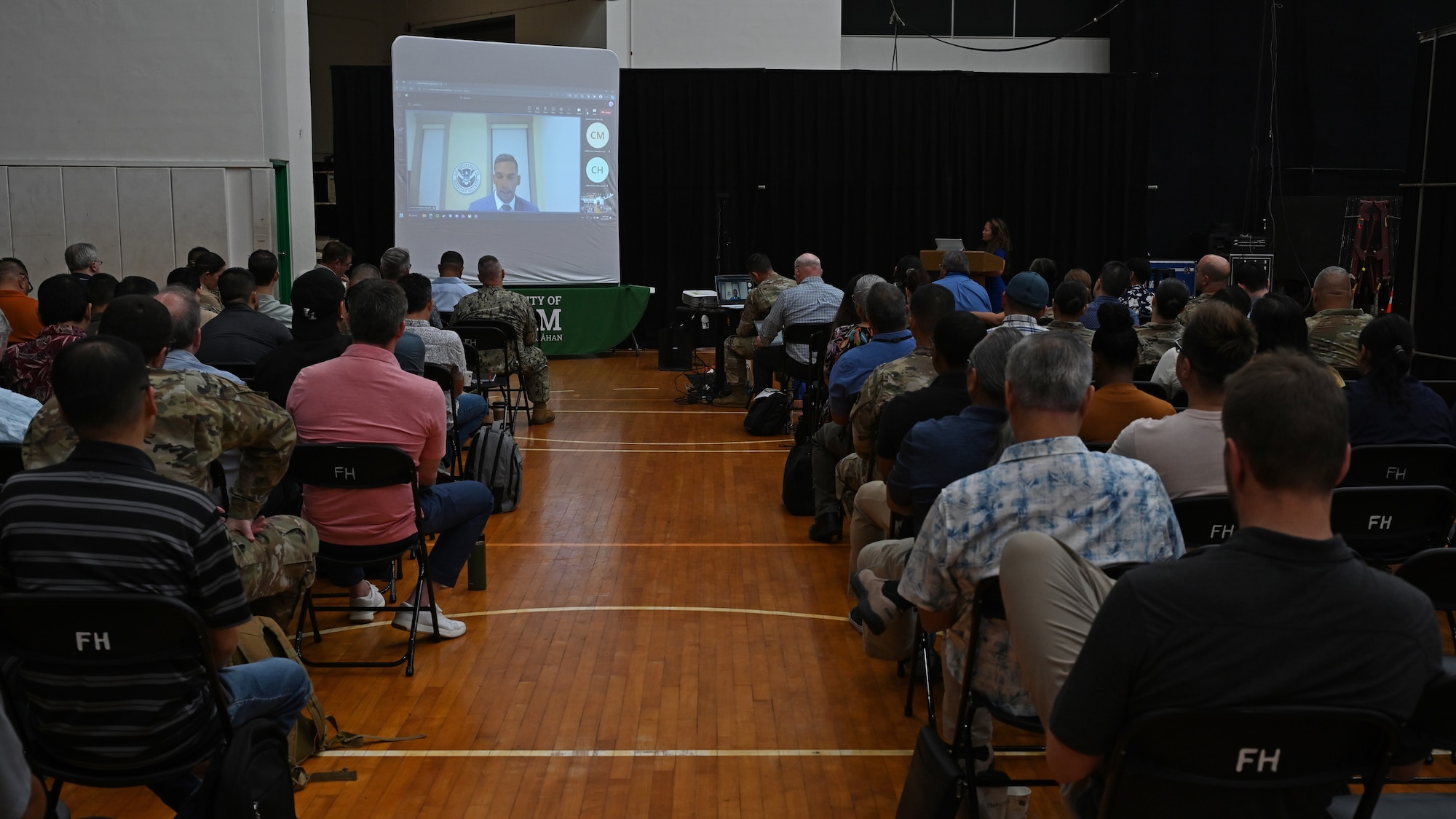 A group of civilians sit on chairs and listen to Iranga Kahangama, assistant secretary of Homeland Security, on the projector screen during the Central Pacific Cybersecurity Summit at the University of Guam.