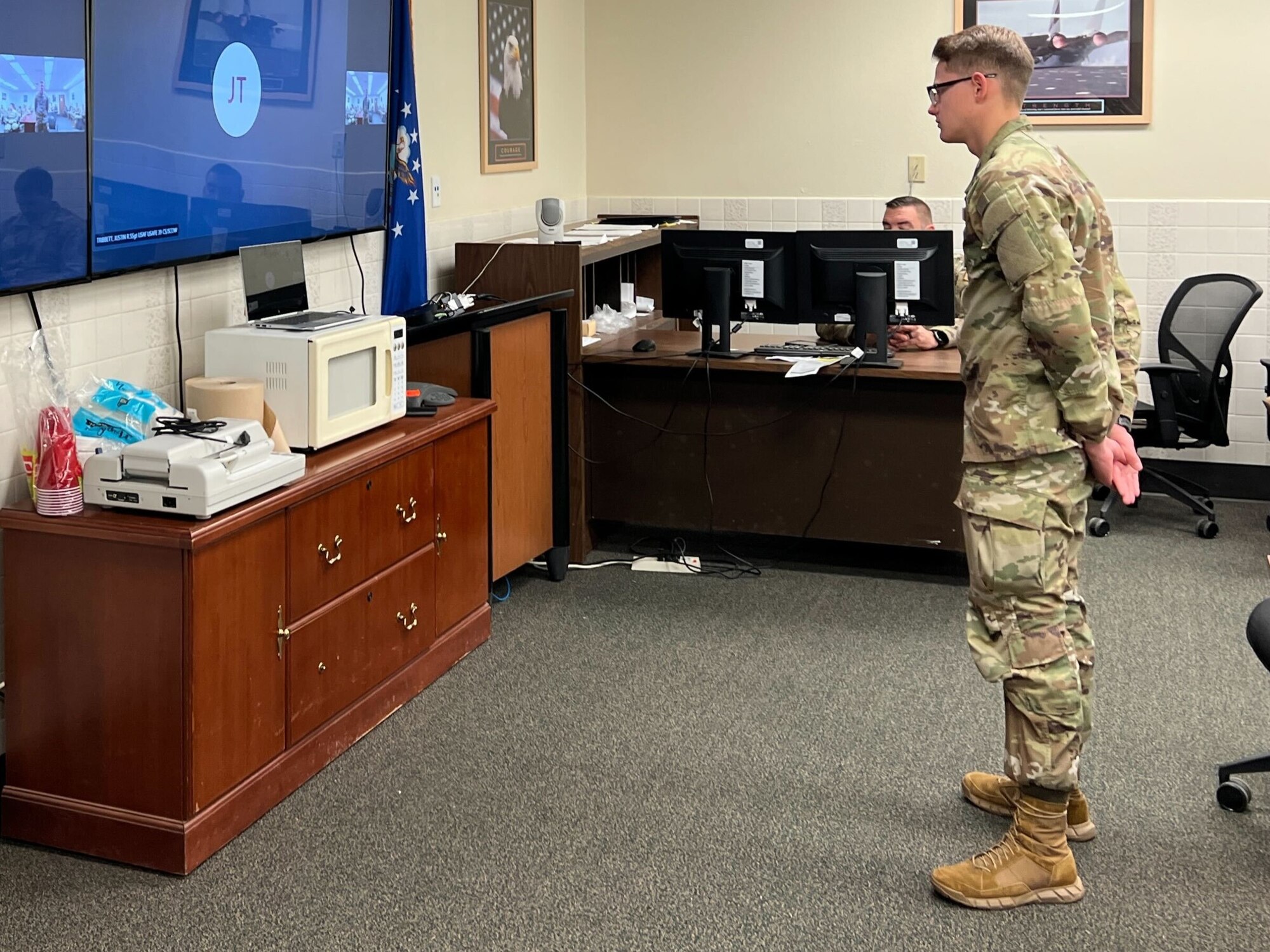 Airmen sit in a conference room. One Airman stands in front of a screen.