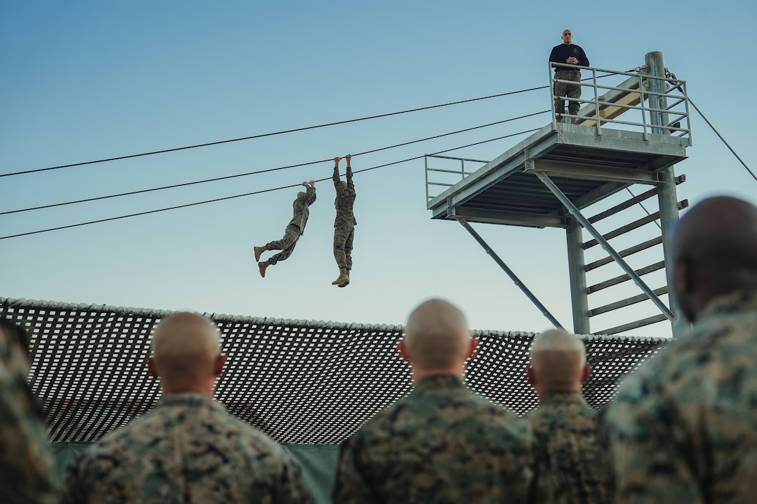 Two Marine Corps recruits hold on to a rope tied between two platforms.