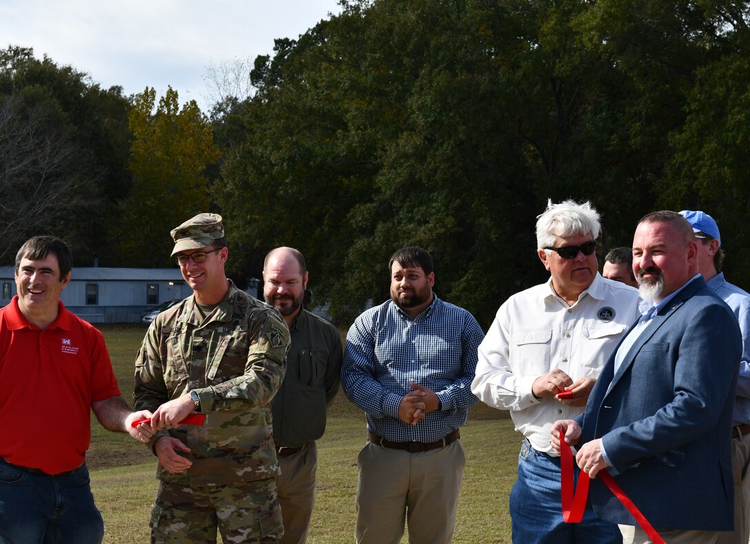 People in business casual clothes and a man in a U.S. Army Colonel's uniform hold pieces of a bright red ribbon in front of a forest.