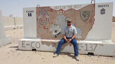 Man with hat and sunglasses sits in front of sign in desert area.
