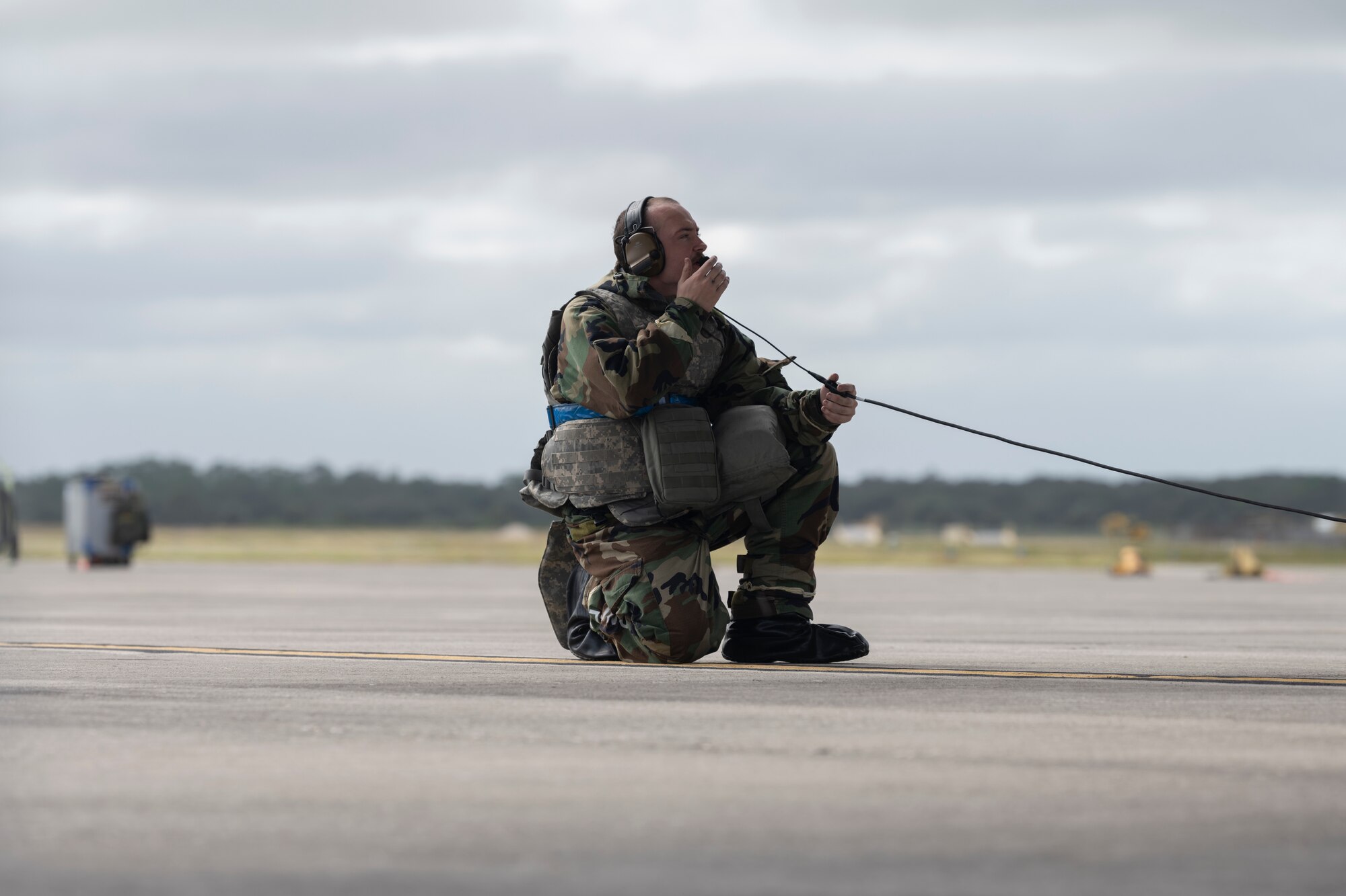A photo of a man in MOPP gear kneeling down on his knee holding his hand to his microphone on his head.