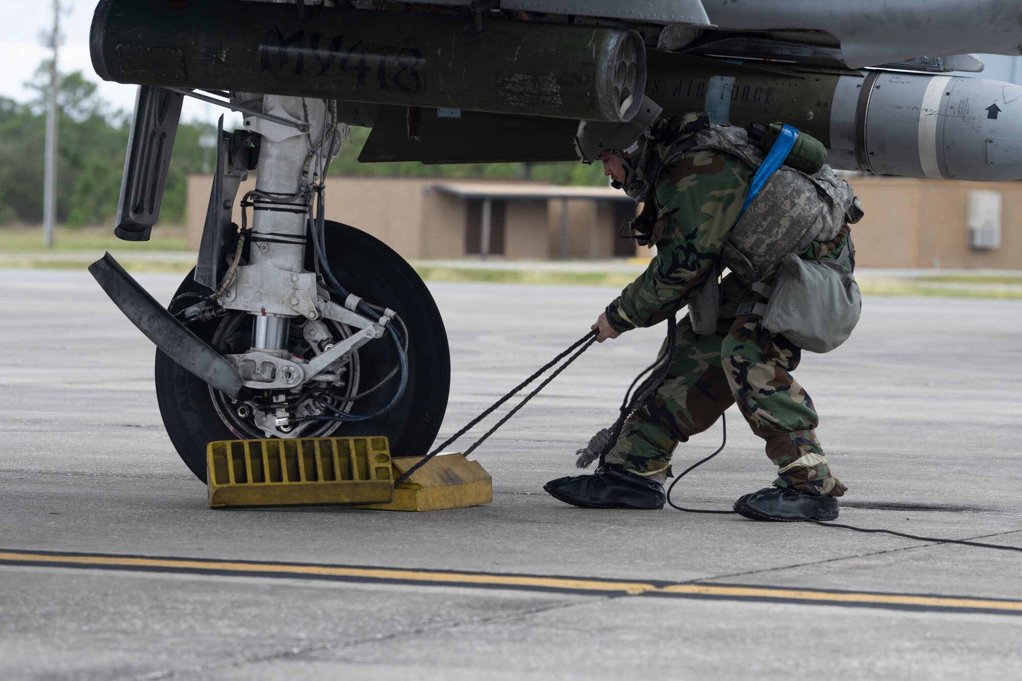 A photo of a person crouching under an A-10C Thunderbolt II, dragging two yellow chocks from the wheel.