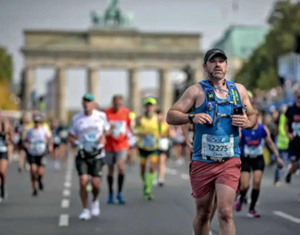 Chris Allen, in focus in the foreground wearing running gear and a racing number, with other runners and Berlin landmarks in the background