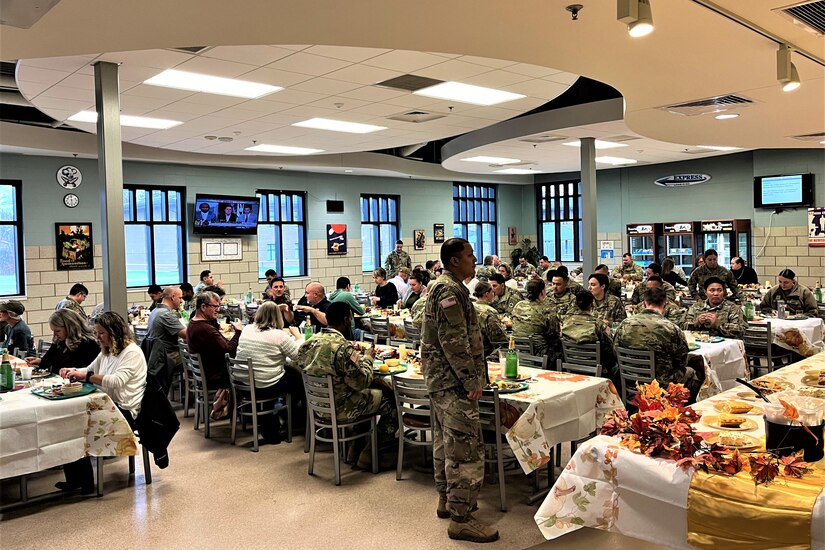 A large group of people, including civilians and uniformed service members, sit and eat at several tables in a cafeteria.