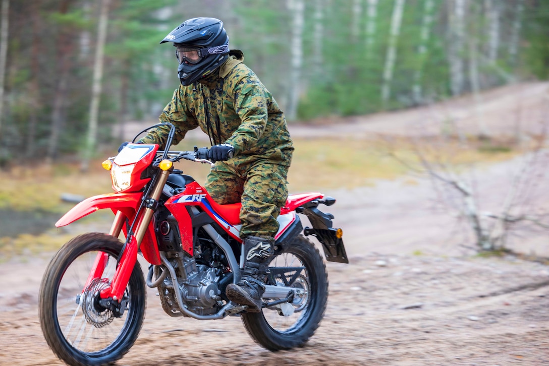 A uniformed sailor rides a dirt bike through a wooded area during daylight.