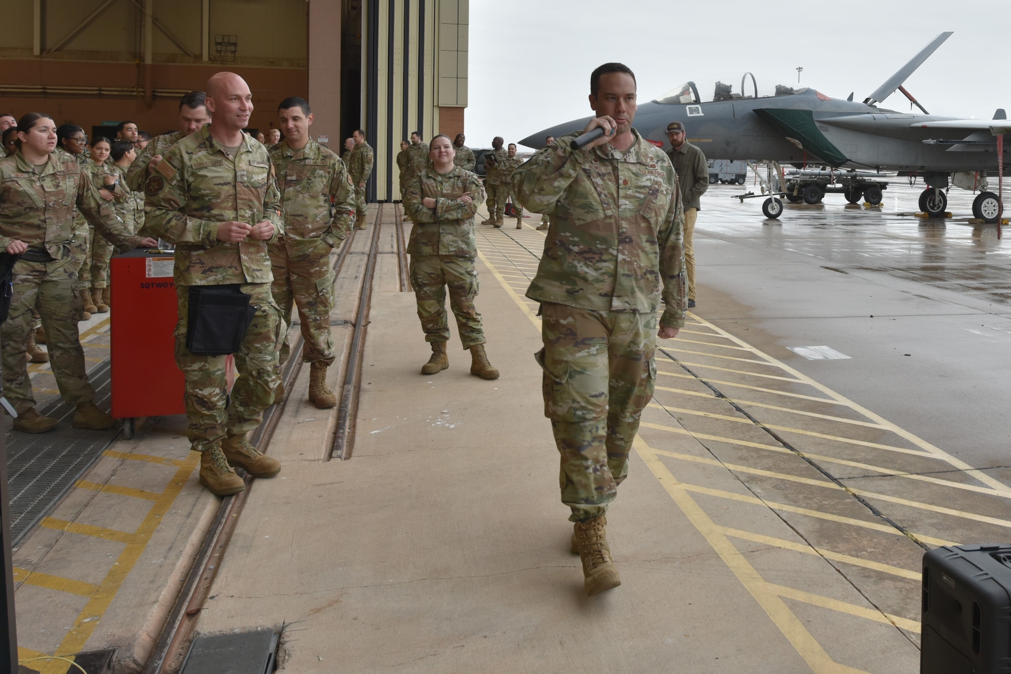 An Air Force officer walks with a microphone as Airmen look on.