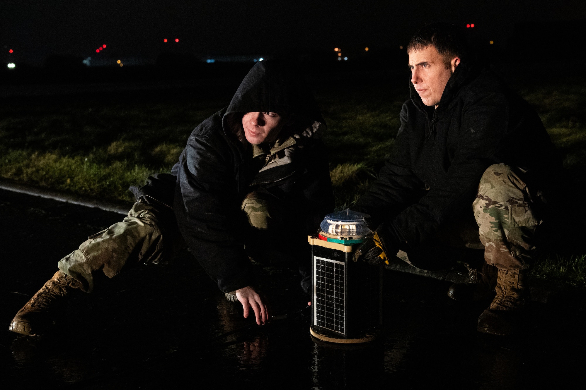 Two Airmen crouch down to shift a light on the ground at night