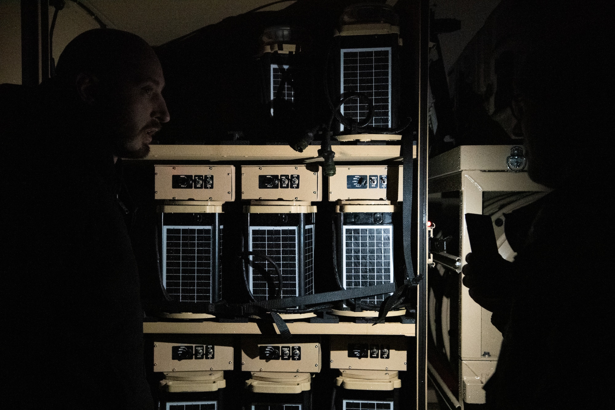 Two Airmen look at runway lights on shelves in a trailer illuminated by a flashlight