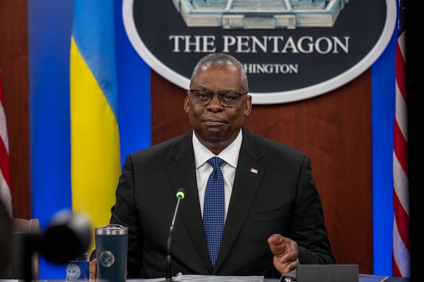 A person in a suit is seated at a table with U.S. and Ukrainian flags in the background.