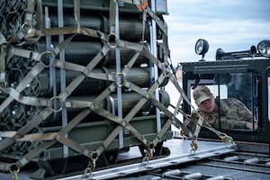 A service member in uniform operates an equipment loader loaded with a pallet of ammunition.