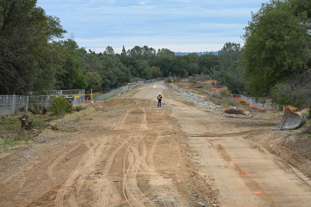 A man in construction gear stands in the middle of an earthen dike. The camera is looking down the length of the dike far away from the man.