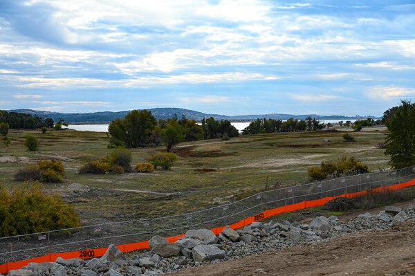 A view of a lake with an earthen dike in the foreground, protected by a chain link fence, and the blue sky with clouds