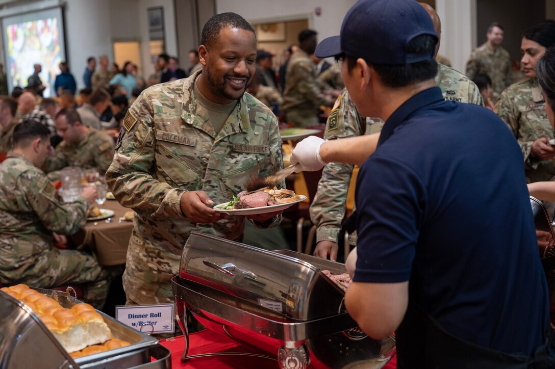 A service member smiles as they receive food for a Thanksgiving dinner as other service members are seated in the background.