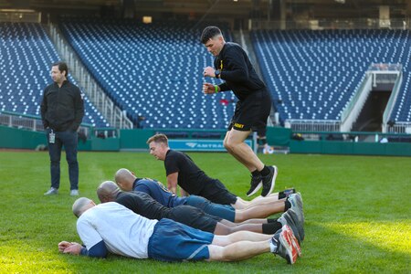 Service members are jumping over each other as others lay flat on the green grass as part of a workout routine.