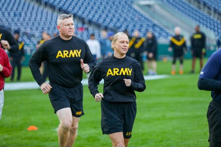 A man and a woman are jogging on green grass. Both are wearing black workout gear that has the word "Army" in large yellow letters.