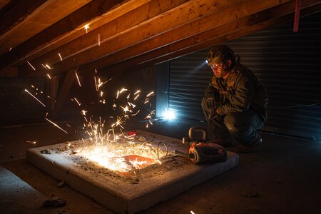 A Marine is squatting in an attic watching another service member cut through a large piece of metal. The cutting is causing dozens of bright yellow sparks to fly into the air.