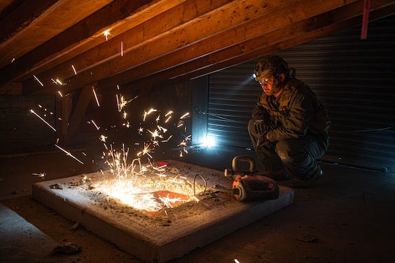 A Marine is squatting in an attic watching another service member cut through a large piece of metal. The cutting is causing dozens of bright yellow sparks to fly into the air.