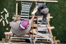 A Marine hands a Soldier a plank of wood as they are climbing on ladders above a green grassy area.