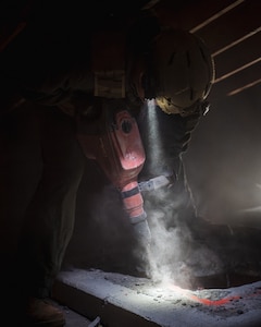 A service member in a dark room uses a red jackhammer to break through cement. There is a bright light attached to his helmet that is illuminating the work area.