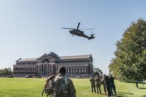 Members of Joint Task Force-National Capital Region stand on the lawn outside a large brick building with a greenish dome in the middle. A helicopter is descending to the lawn.