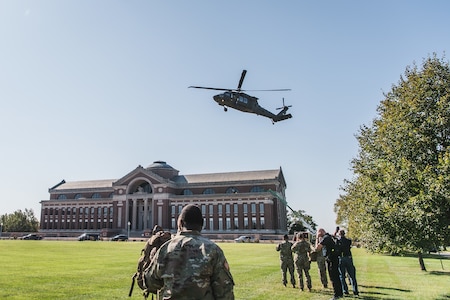 Members of Joint Task Force-National Capital Region stand on the lawn outside a large brick building with a greenish dome in the middle. A helicopter is descending to the lawn.