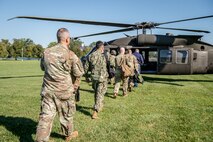 Members of Joint Task Force-National Capital Region walk in a line toward a helicopter, which is parked on green grass.