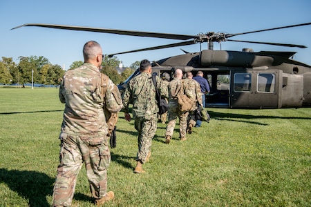 Members of Joint Task Force-National Capital Region walk in a line toward a helicopter, which is parked on green grass.