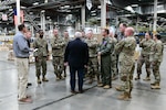 Photo is of a group of people, some in military uniform, standing in a small aisle in a warehouse.