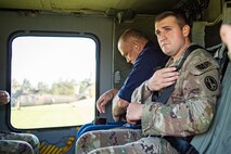 An Army service member in fatigues adjusts his seat belt while seated inside a helicopter next to a man dress in blue, who is also adjusting his seat belt. Through the window is a helicopter on the ground in the distance