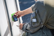 Member of the FBI is holding up an electronic device to a bright green piece of tap that is affixed to a beige container