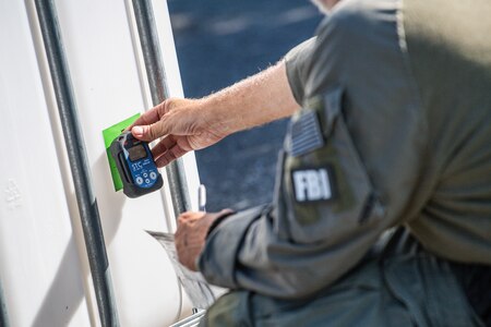 Member of the FBI is holding up an electronic device to a bright green piece of tap that is affixed to a beige container