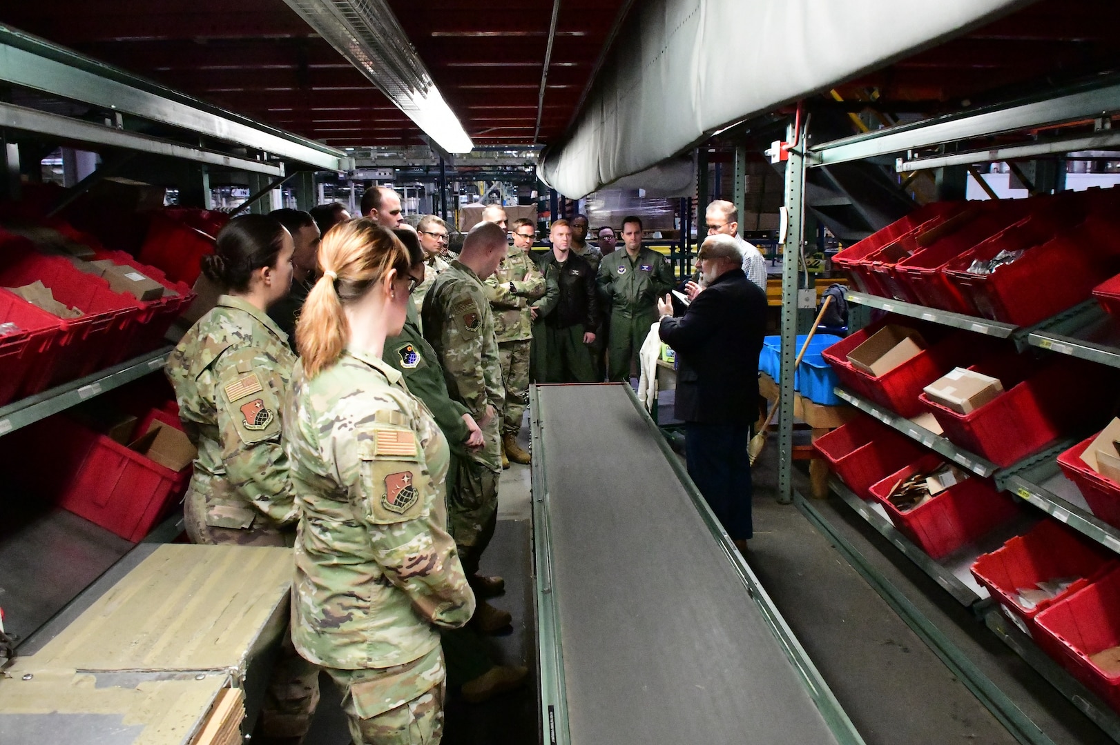 Photo is of a group of people, some in military uniform, standing in a small aisle in a warehouse.