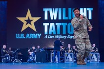 An Army Soldier dressed in green fatigues is singing into a micro phone. In the background, a band is performing while wearing dark blue ceremonial uniforms, and above them on a screen is the U.S. Army logo with yellow star and the words Twilight Tattoo, a live military experience